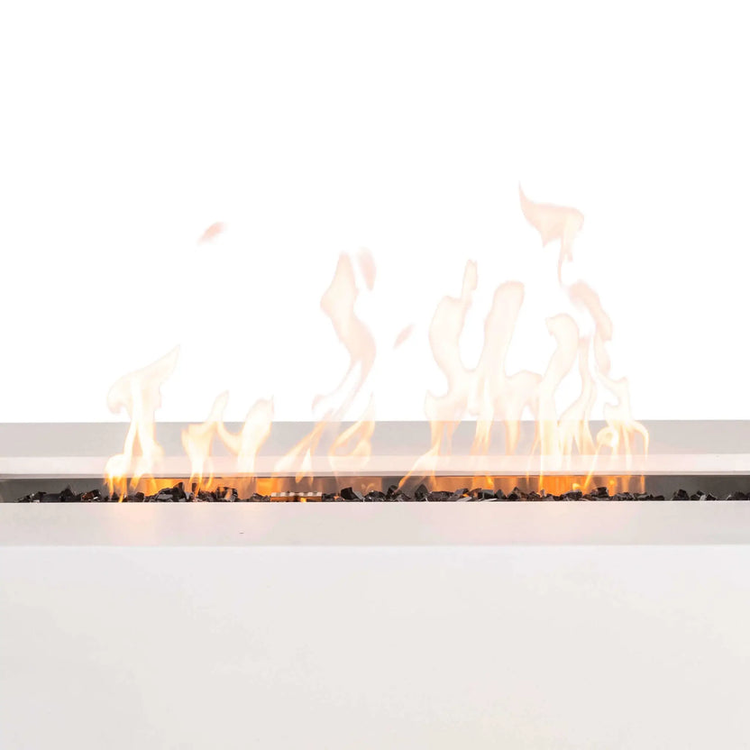 Close-up of the Flame Havens Refuge Series 56 x 30 x 24-inch rectangular white propane gas fire pit burning with tall yellow-orange flames across tempered black fire glass, highlighting the linear burner, stainless steel pan, central ignition cage, and wide white GFRC concrete ledge.
