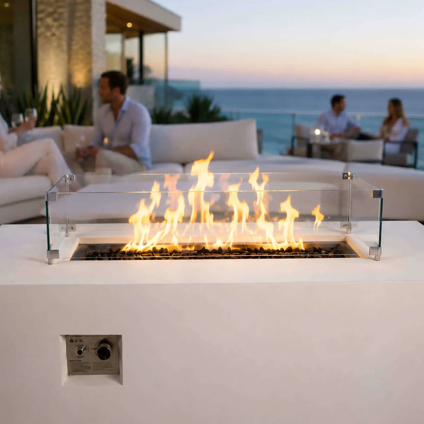 Lifestyle view of the Flame Havens Refuge Series 56 x 30 x 24-inch rectangular white propane gas fire pit burning over tempered black fire glass with a clear wind guard, creating a warm focal point for guests relaxing on a modern coastal patio at sunset.