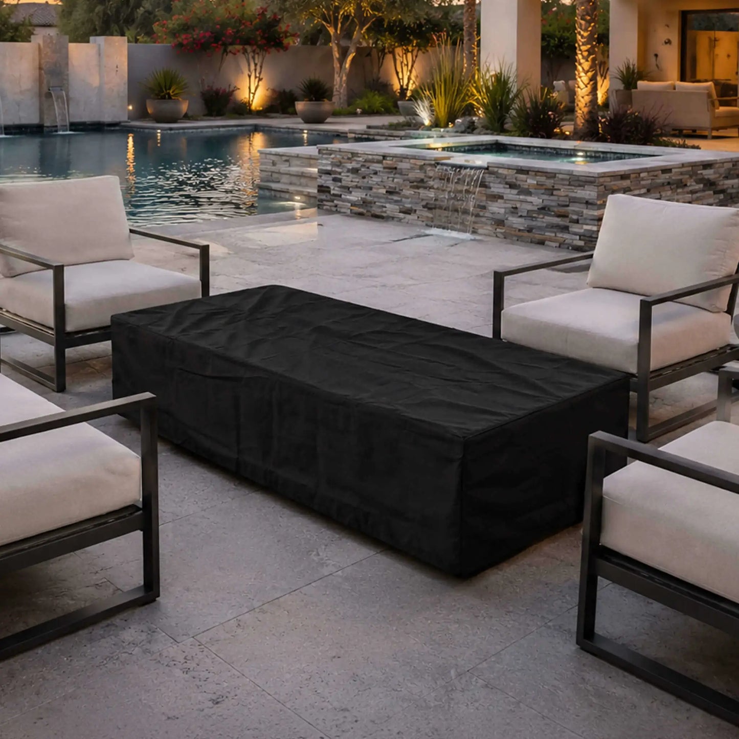 Flame Havens Refuge Series 68 x 30 x 15-inch rectangular fire pit protected by a fitted black canvas fabric cover. The covered fire pit sits between cushioned patio chairs beside a luxury swimming pool and raised spa with a stacked-stone waterfall in a warmly lit outdoor living space.