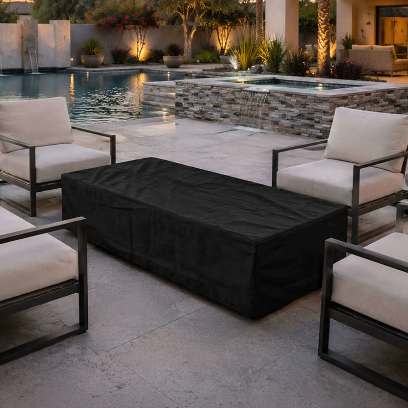 Flame Havens Refuge Series 68 x 30 x 15-inch rectangular fire pit protected by a fitted black canvas fabric cover. The covered fire pit sits between cushioned patio chairs beside a luxury swimming pool and raised spa with a stacked-stone waterfall in a warmly lit outdoor living space.