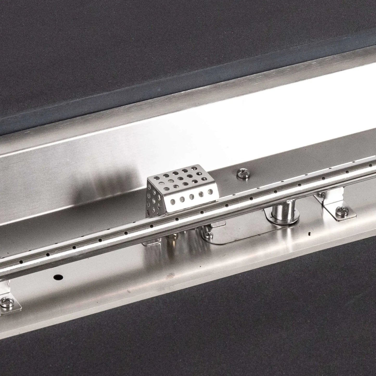 Tight detail of the stainless steel burner and CSA ignition components inside the Flame Havens Refuge Series 68 x 30 x 15-inch rectangular natural gas fire pit. The image shows the linear burner ports, perforated ignition housing, hardware, supports, and brushed metal pan surrounded by black GFRC concrete.