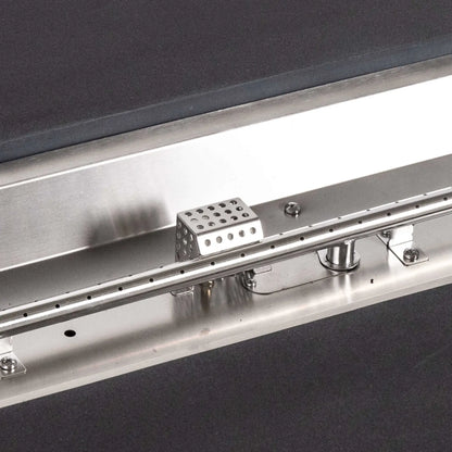 Tight detail of the stainless steel burner and CSA ignition components inside the Flame Havens Refuge Series 68 x 30 x 15-inch rectangular natural gas fire pit. The image shows the linear burner ports, perforated ignition housing, hardware, supports, and brushed metal pan surrounded by black GFRC concrete.