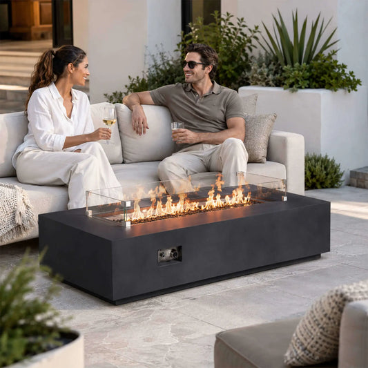 Couple relaxing with drinks beside a lit Flame Havens Refuge Series 68 x 30 x 15-inch rectangular natural gas fire pit in black GFRC concrete. Flames rise through tempered black fire glass behind a clear glass wind guard, while the front control panel remains accessible in the modern outdoor patio setting.