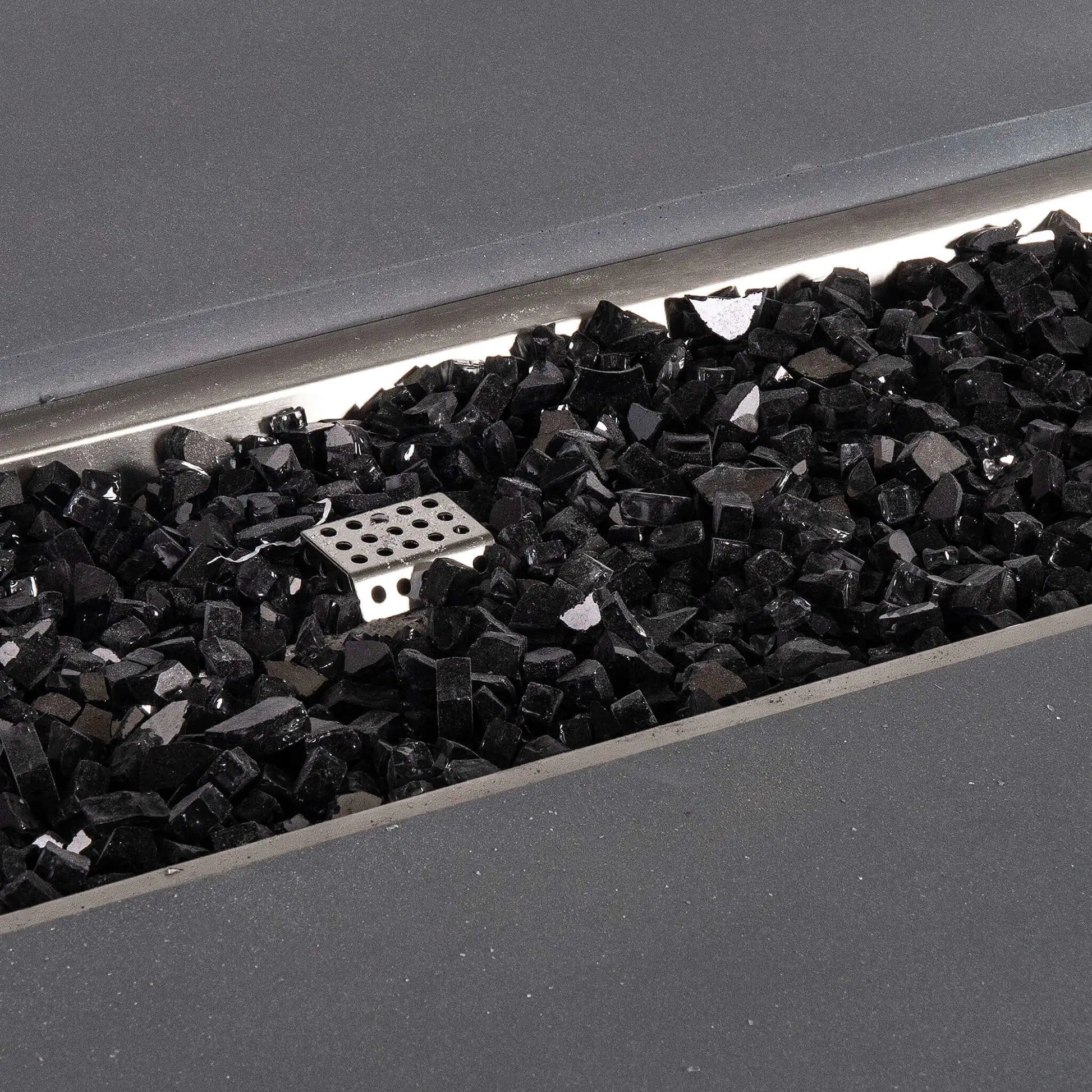 Detailed close-up of reflective black fire glass inside the Flame Havens Refuge Series 68 x 30 x 15-inch rectangular natural gray GFRC concrete fire pit. The glossy broken glass pieces surround the stainless steel ignition cage within the recessed natural gas burner pan.