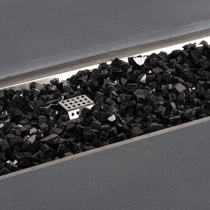 Detailed close-up of reflective black fire glass inside the Flame Havens Refuge Series 68 x 30 x 15-inch rectangular natural gray GFRC concrete fire pit. The glossy broken glass pieces surround the stainless steel ignition cage within the recessed natural gas burner pan.