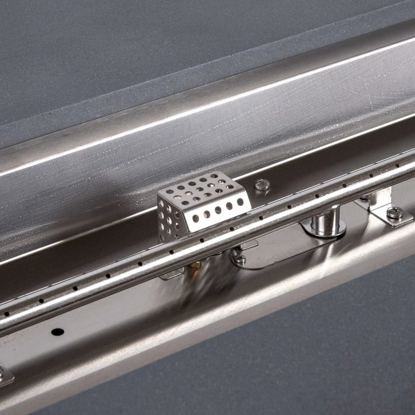 Tight close-up of the Flame Havens Refuge Series stainless steel natural gas burner and CSA ignition assembly. The image clearly shows the linear burner ports, perforated ignition cage, mounting brackets, fasteners, and reflective burner pan installed inside the 68 x 30 x 15-inch natural gray GFRC fire pit.