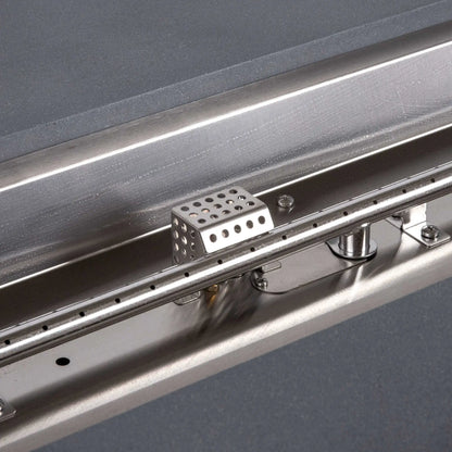 Tight close-up of the Flame Havens Refuge Series stainless steel natural gas burner and CSA ignition assembly. The image clearly shows the linear burner ports, perforated ignition cage, mounting brackets, fasteners, and reflective burner pan installed inside the 68 x 30 x 15-inch natural gray GFRC fire pit.