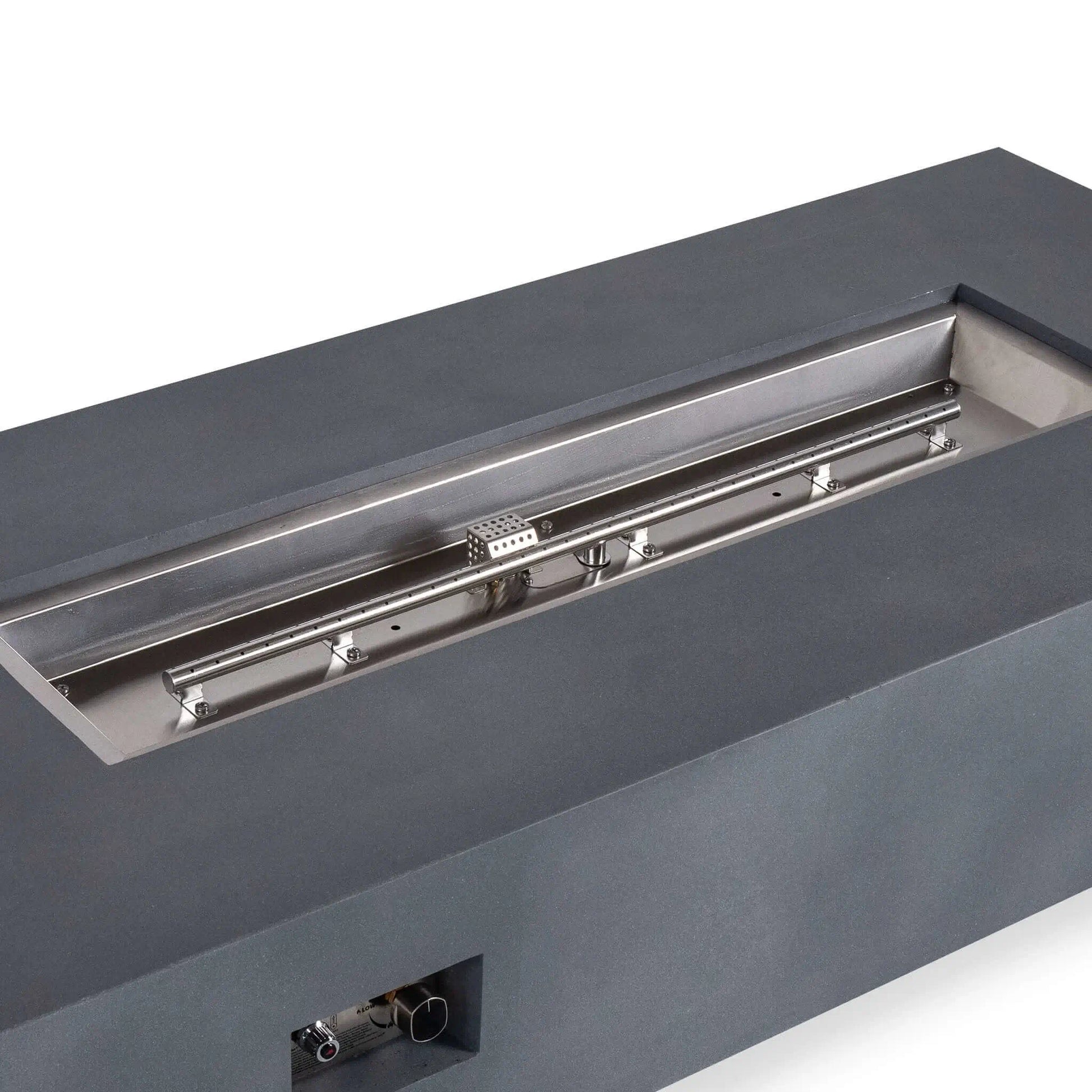 Close angled view of the Flame Havens Refuge Series 68 x 30 x 15-inch natural gray GFRC fire pit with the fire media removed. The image highlights the stainless steel burner pan, linear natural gas burner, ignition cage, mounting hardware, and recessed front control panel.