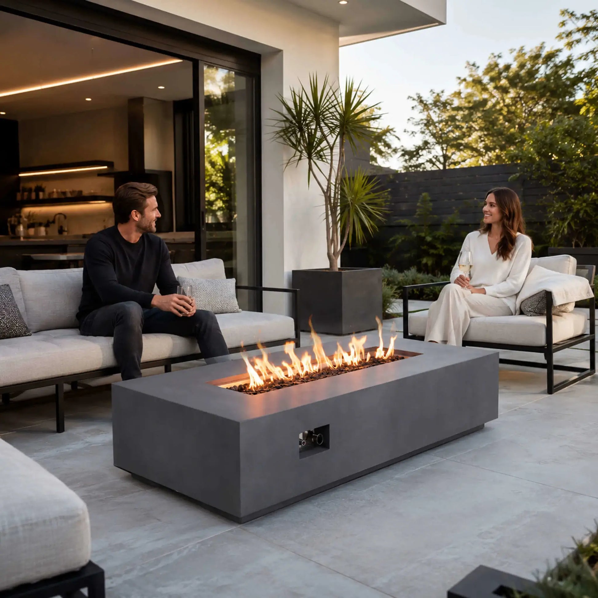 A couple relaxes with drinks around the Flame Havens Refuge Series 68 x 30 x 15-inch rectangular natural gas fire pit on a modern covered patio. Flames glow above tempered black fire glass in the natural gray GFRC fire pit, surrounded by white outdoor seating, contemporary landscaping, and an open-air kitchen.