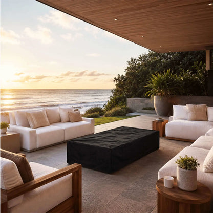 Flame Havens Refuge Series 68 x 30 x 15-inch white GFRC rectangular natural gas fire pit protected by a fitted black canvas fabric cover. The covered fire pit is centered among white outdoor sofas on a luxurious oceanfront patio at sunset, beneath a warm wood ceiling with coastal landscaping nearby.