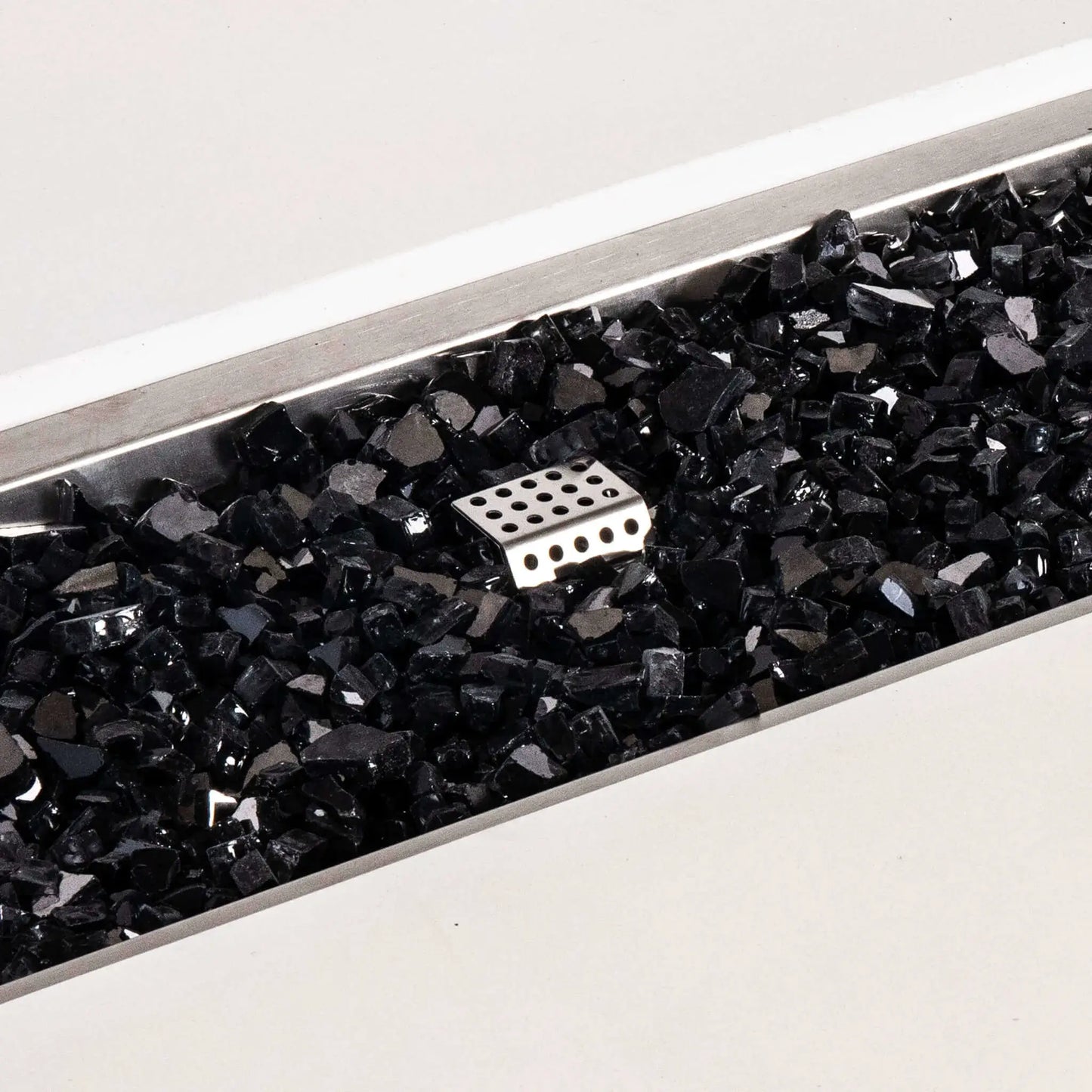 Close-up of the Flame Havens Refuge Series white GFRC concrete fire pit showing glossy black tempered fire glass inside the rectangular stainless steel burner pan. A perforated metal ignition cage rises through the reflective glass media, highlighting the natural gas fire pit’s burner components and clean finish.