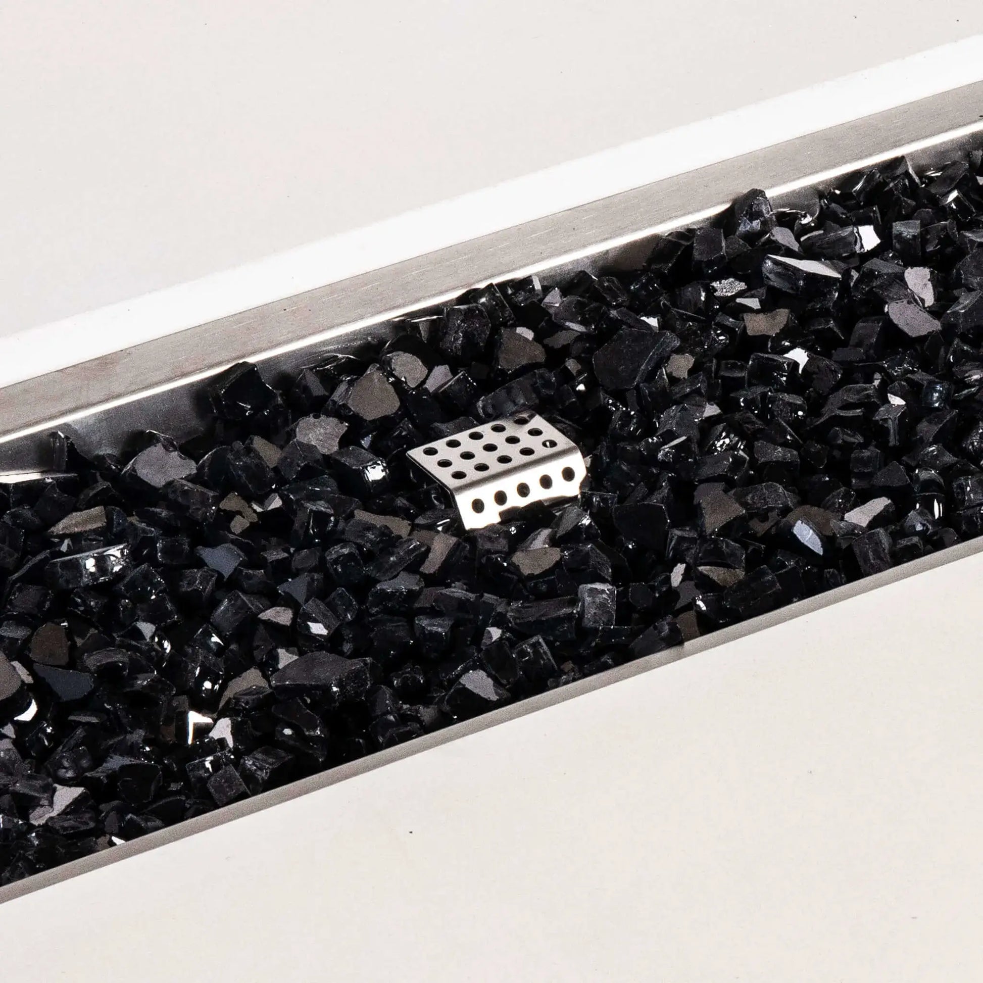 Close-up of the Flame Havens Refuge Series white GFRC concrete fire pit showing glossy black tempered fire glass inside the rectangular stainless steel burner pan. A perforated metal ignition cage rises through the reflective glass media, highlighting the natural gas fire pit’s burner components and clean finish.