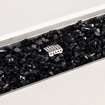 Close-up of the Flame Havens Refuge Series white GFRC concrete fire pit showing glossy black tempered fire glass inside the rectangular stainless steel burner pan. A perforated metal ignition cage rises through the reflective glass media, highlighting the natural gas fire pit’s burner components and clean finish.