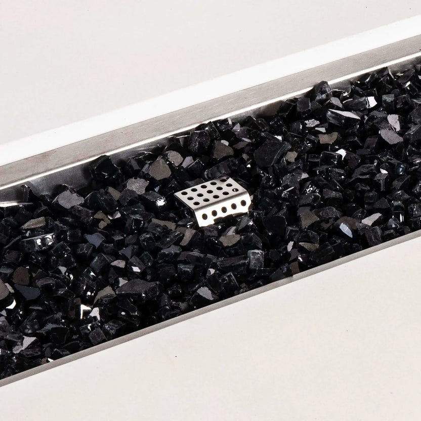 Close-up of the Flame Havens Refuge Series white GFRC concrete fire pit showing glossy black tempered fire glass inside the rectangular stainless steel burner pan. A perforated metal ignition cage rises through the reflective glass media, highlighting the natural gas fire pit’s burner components and clean finish.