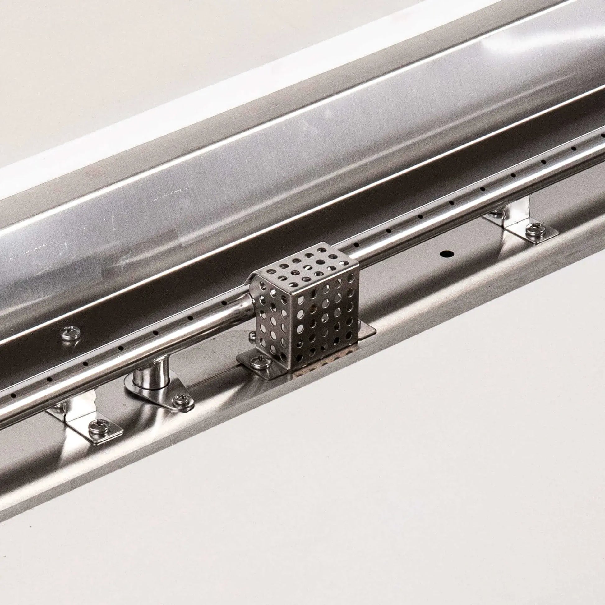 Detailed close-up of the Flame Havens Refuge Series natural gas fire pit’s stainless steel burner and CSA ignition assembly. The image highlights the linear burner ports, polished mounting hardware, ignition components and perforated protective cage installed inside the white 68 x 30-inch rectangular GFRC fire pit.