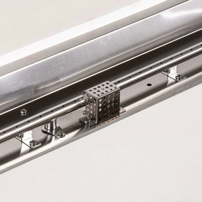 Detailed close-up of the Flame Havens Refuge Series natural gas fire pit’s stainless steel burner and CSA ignition assembly. The image highlights the linear burner ports, polished mounting hardware, ignition components and perforated protective cage installed inside the white 68 x 30-inch rectangular GFRC fire pit.
