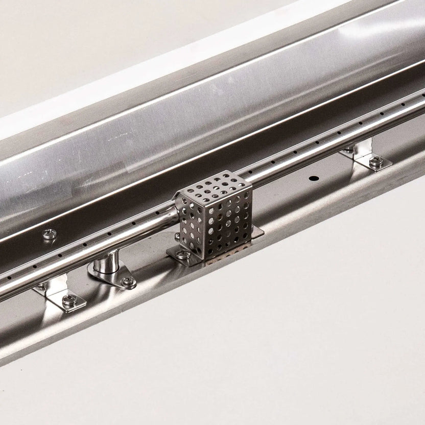 Detailed close-up of the Flame Havens Refuge Series natural gas fire pit’s stainless steel burner and CSA ignition assembly. The image highlights the linear burner ports, polished mounting hardware, ignition components and perforated protective cage installed inside the white 68 x 30-inch rectangular GFRC fire pit.