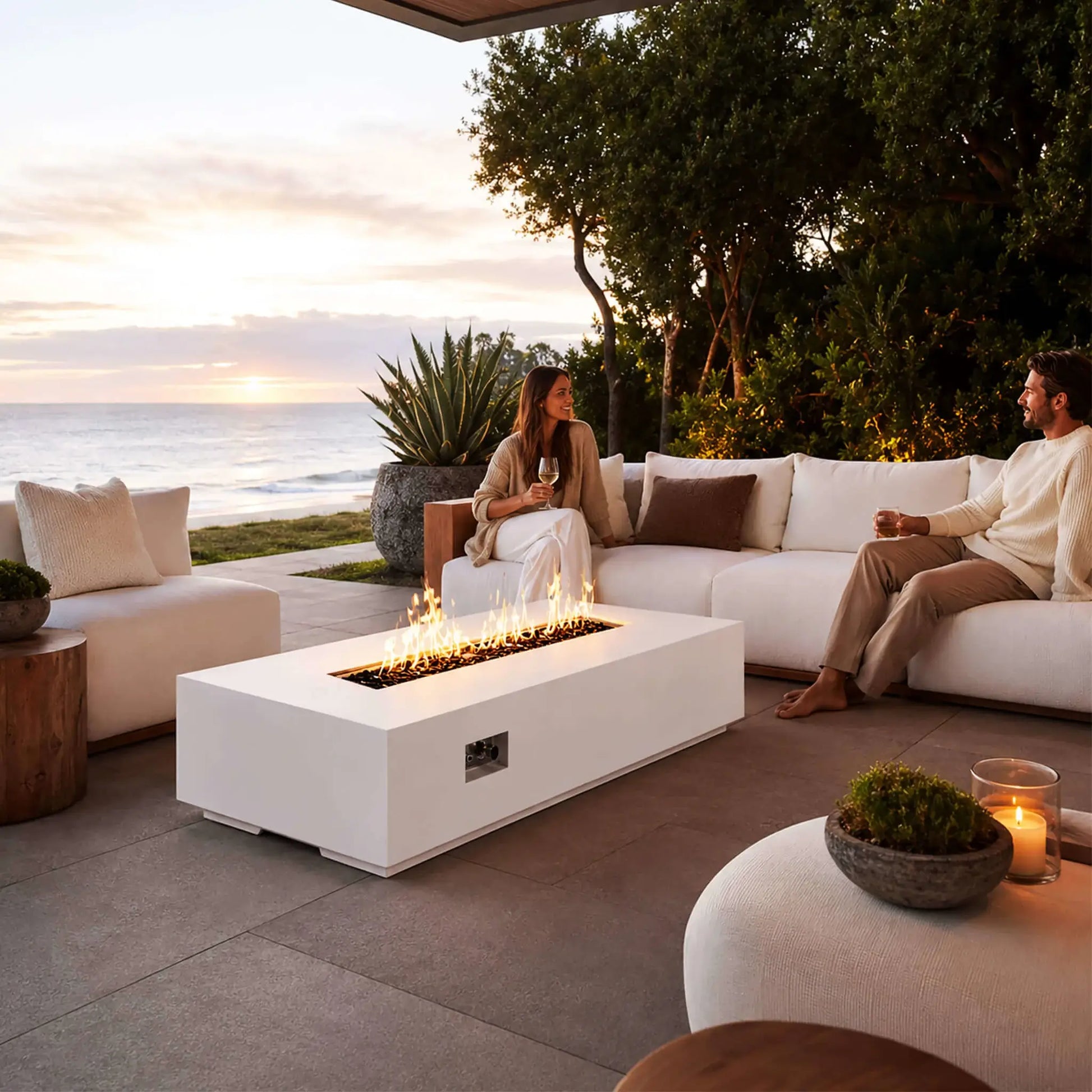 Flame Havens Refuge Series 68 x 30 x 15-inch white GFRC rectangular natural gas fire pit glowing on a coastal patio at sunset. Flames rise from tempered black fire glass, and the recessed controls are visible on the front. A couple relaxes with drinks on white outdoor seating overlooking the ocean.