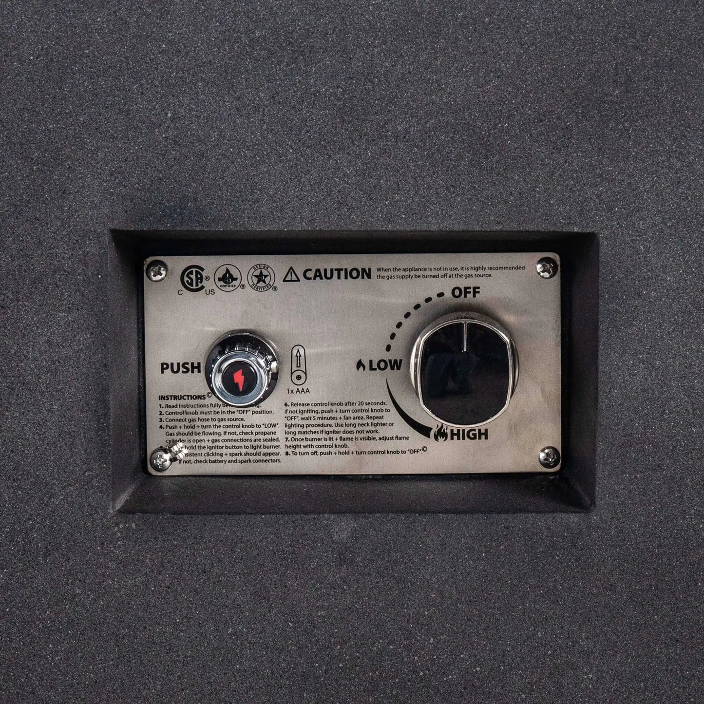 Close-up of the recessed CSA ignition controls on the Flame Havens Refuge Series 68 x 30 x 15-inch rectangular natural gas fire pit. The stainless steel panel includes a push-button spark igniter, AAA battery indicator, operating instructions, safety markings, and a large flame-control knob labeled off, low, and high.