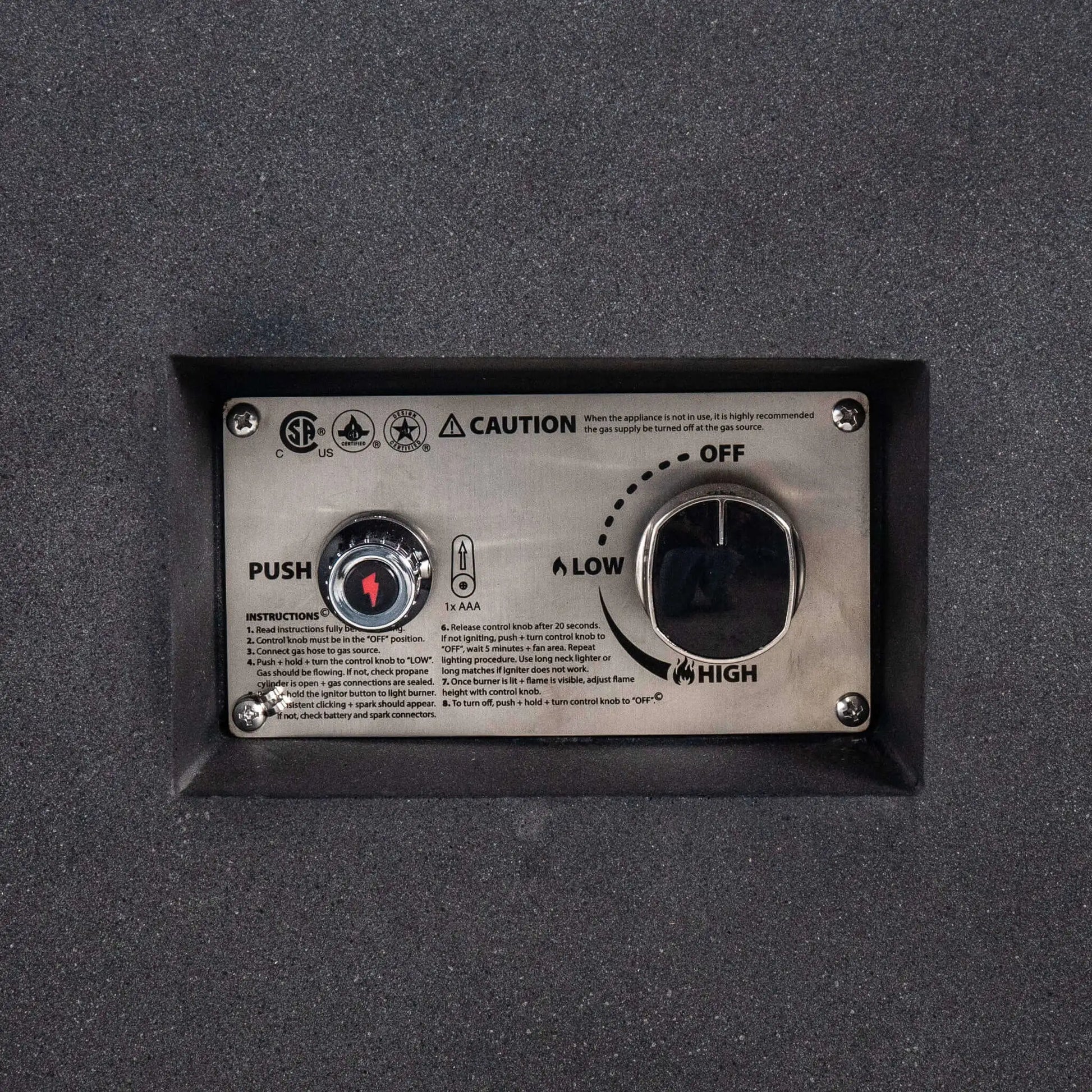 Close-up of the recessed CSA ignition controls on the Flame Havens Refuge Series 68 x 30 x 15-inch rectangular natural gas fire pit. The stainless steel panel includes a push-button spark igniter, AAA battery indicator, operating instructions, safety markings, and a large flame-control knob labeled off, low, and high.
