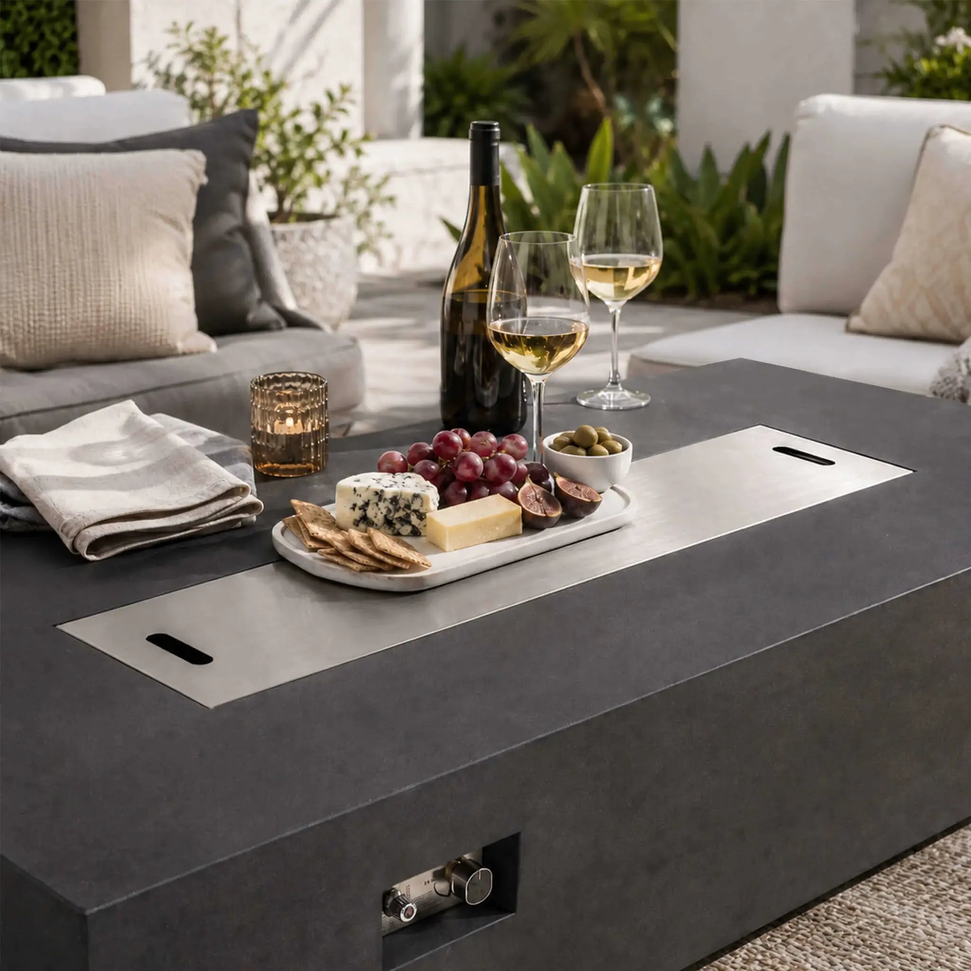 Flame Havens Refuge Series 68 x 30 x 15-inch black GFRC natural gas fire pit styled for outdoor entertaining with its stainless steel burner cover installed. The covered surface holds wine, glasses, grapes, cheese, crackers, figs, olives, and a candle between cushioned patio sofas.