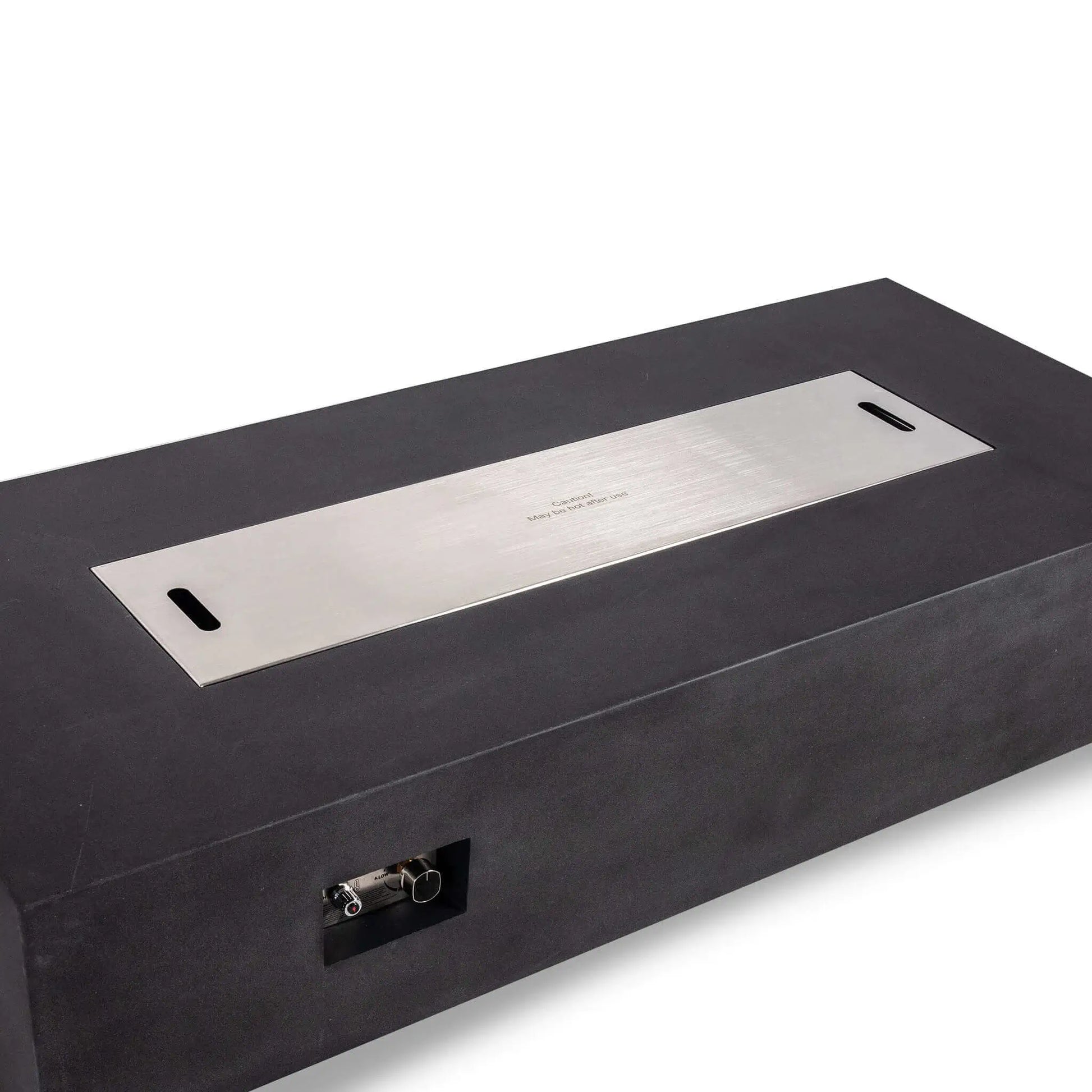 Angled view of a Flame Havens Refuge Series 68 x 30 x 15-inch rectangular natural gas fire pit in black GFRC concrete. A flush-fitting brushed stainless steel cover conceals the burner opening and includes two recessed lifting slots and a centered caution marking. The front ignition controls remain visible.