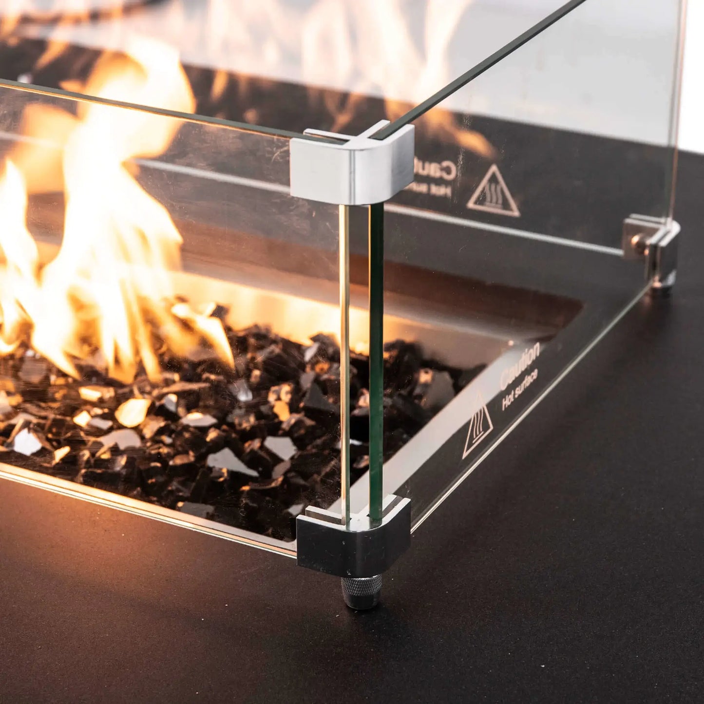 Close-up of the tempered glass wind guard surrounding active flames and reflective black fire glass on a Flame Havens Refuge Series 68 x 30 x 15-inch natural gas fire pit. The image highlights the clear glass panels, polished metal corner brackets, adjustable support foot, and warm flame reflections.