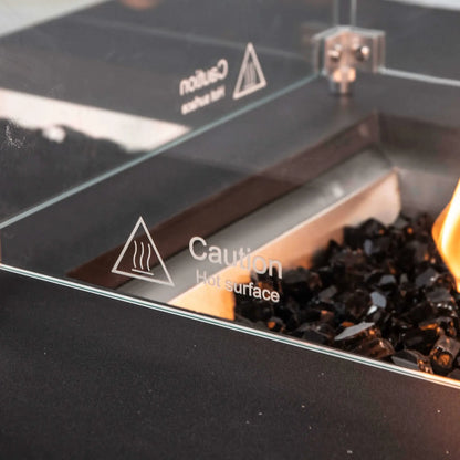 Detailed close-up of the tempered glass wind guard on a Flame Havens Refuge Series 68 x 30 x 15-inch black GFRC natural gas fire pit. A visible “Caution Hot Surface” marking appears on the clear glass above reflective black fire glass, with an orange flame glowing along the edge of the burner.