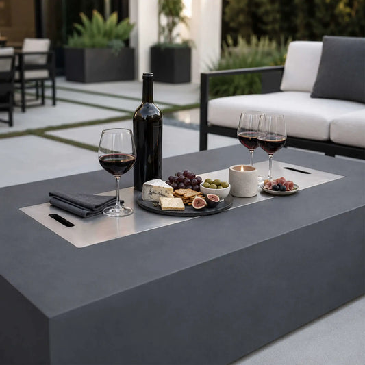 Flame Havens Refuge Series 68 x 30 x 15-inch natural gray GFRC fire pit styled as an outdoor table with the stainless steel burner cover installed. Wine, glasses, cheese, crackers, fruit, olives, and a candle are arranged across the covered burner area on a contemporary patio.