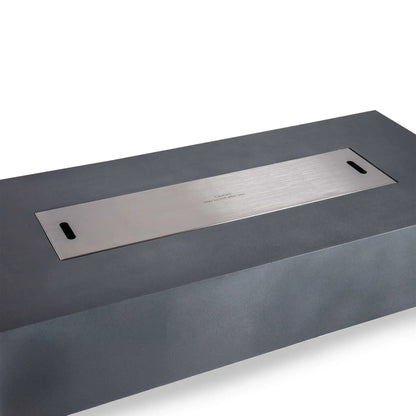 Angled view of the Flame Havens Refuge Series 68 x 30 x 15-inch natural gray GFRC concrete fire pit with its stainless steel burner cover installed. The long brushed-metal cover fits neatly over the natural gas burner opening and includes two hand slots and a centered hot-surface caution message.