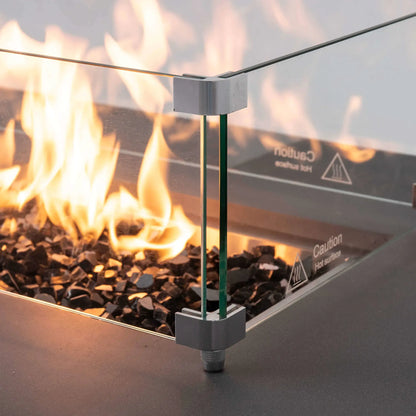 Close-up of the tempered glass wind guard surrounding bright flames and tempered black fire glass on the Flame Havens Refuge Series 68 x 30 x 15-inch natural gas fire pit. The image highlights the clear glass panels, metal corner brackets, adjustable foot, and reflections from the burning linear flame.