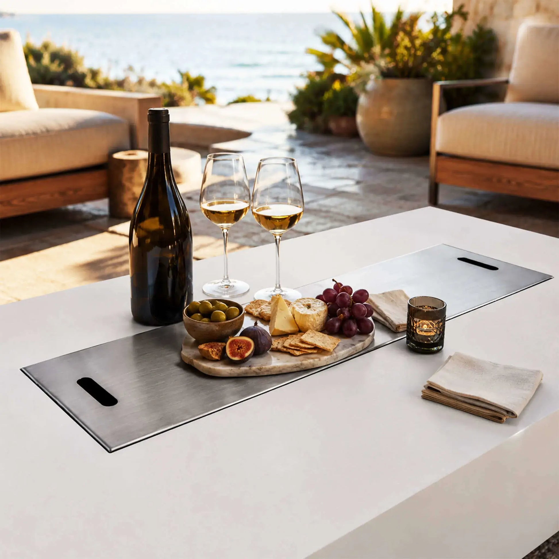 Flame Havens Refuge Series 68 x 30 x 15-inch white GFRC natural gas fire pit with its stainless steel burner cover used as a serving surface. Wine, glasses, cheese, crackers, grapes, figs and olives are arranged on the brushed metal cover in a sunlit oceanfront patio setting.