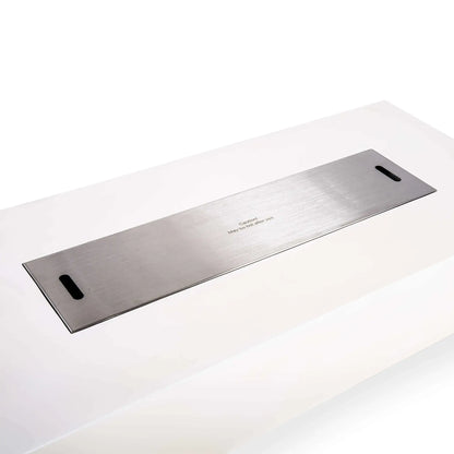 Stainless steel burner cover fitted over the opening of a Flame Havens Refuge Series 68 x 30 x 15-inch rectangular natural gas fire pit in white GFRC concrete. The long brushed metal cover sits flush with the top and includes two handle openings and a centered caution message.