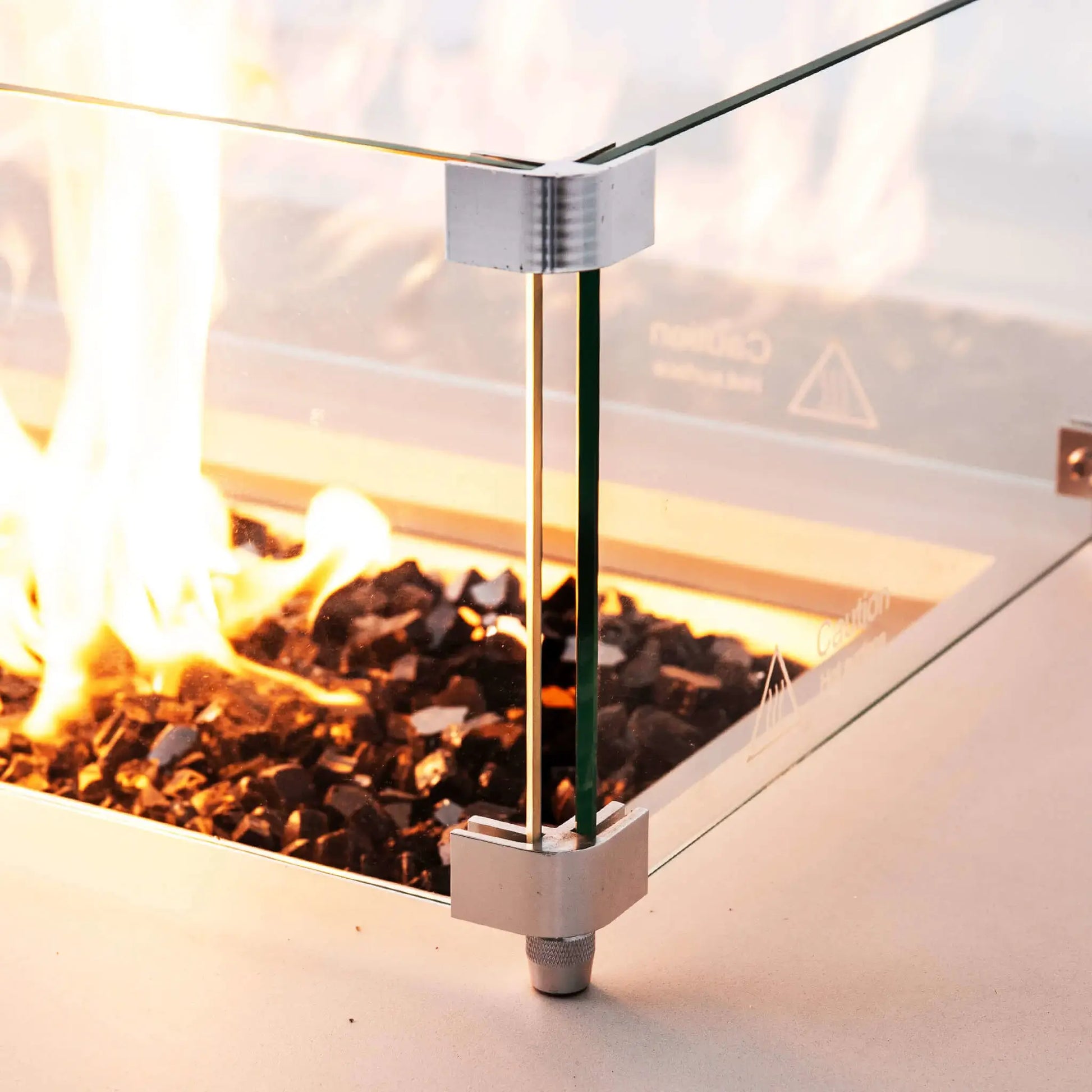 Close-up of the tempered glass wind guard surrounding the lit burner on a Flame Havens Refuge Series 68 x 30 x 15-inch white GFRC natural gas fire pit. Stainless steel corner brackets and an adjustable foot secure the clear glass panels as bright flames rise from reflective tempered black fire glass.