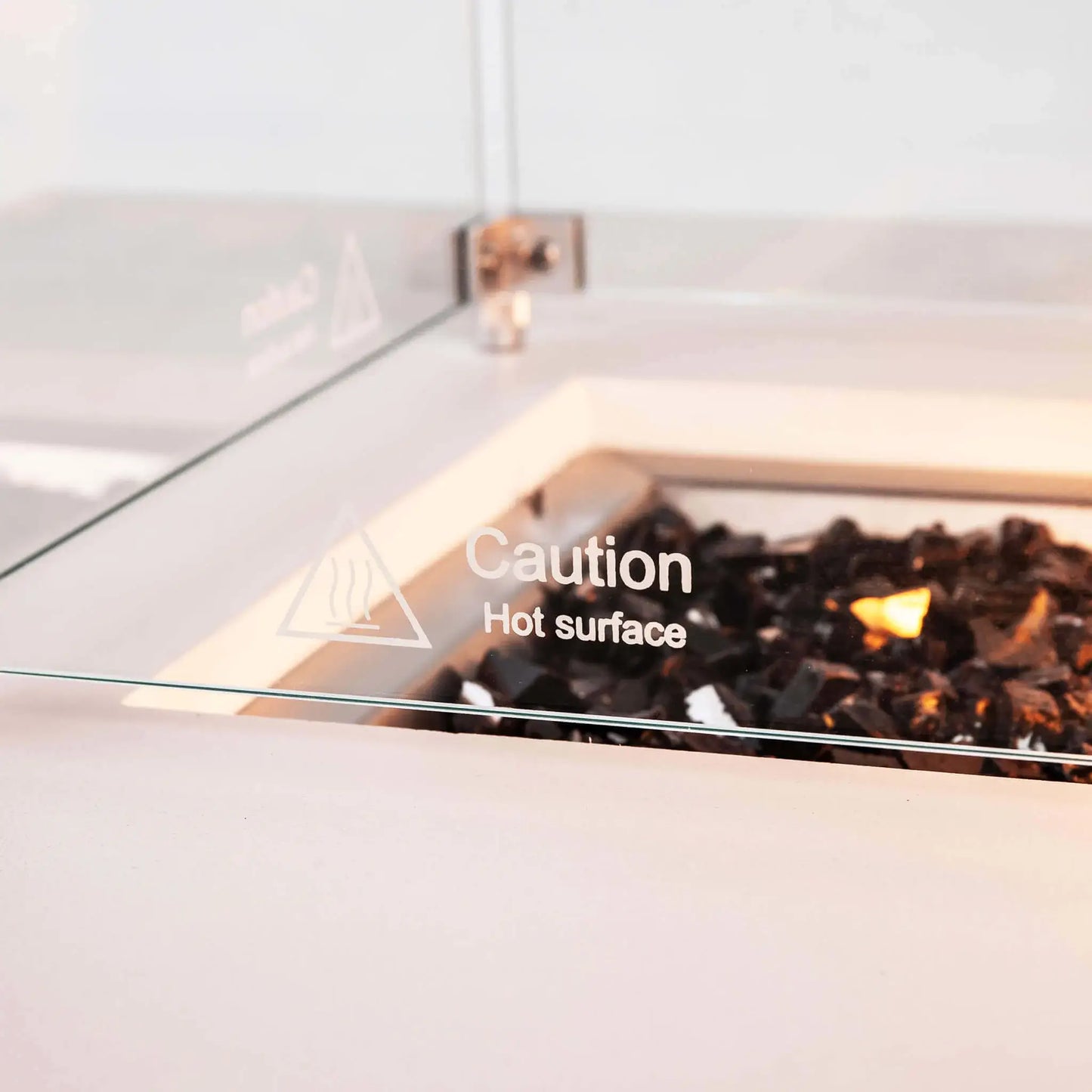 Detailed view of the tempered glass wind guard on a Flame Havens Refuge Series white GFRC rectangular natural gas fire pit. A printed “Caution Hot Surface” warning is visible on the clear glass, with reflective tempered black fire glass and the warm glow of the operating burner behind it.