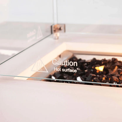 Detailed view of the tempered glass wind guard on a Flame Havens Refuge Series white GFRC rectangular natural gas fire pit. A printed “Caution Hot Surface” warning is visible on the clear glass, with reflective tempered black fire glass and the warm glow of the operating burner behind it.