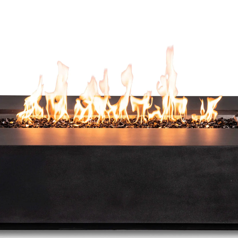Close-up of active natural gas flames stretching across reflective tempered black fire glass in the Flame Havens Refuge Series 68 x 30 x 15-inch rectangular fire pit. The image highlights the varied flame heights, stainless steel burner-pan edge, dark GFRC concrete finish, and concealed-control design.