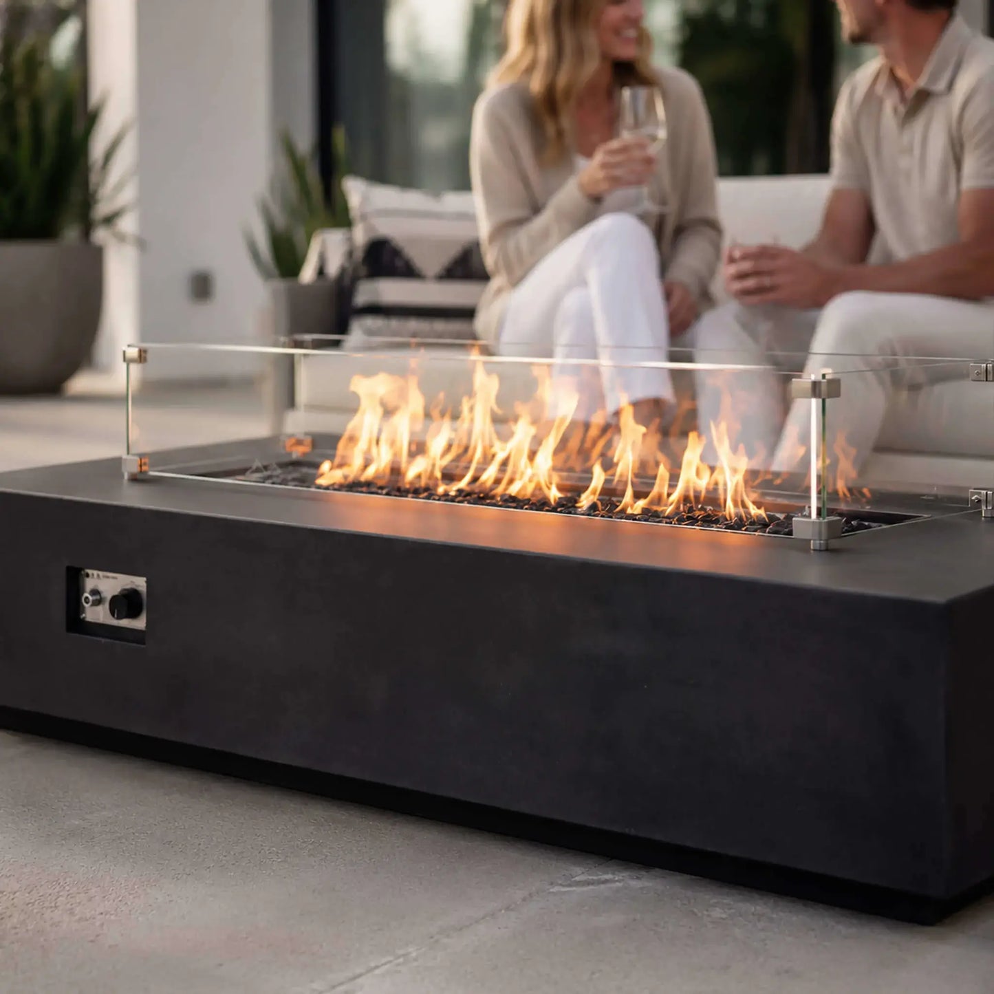 Couple relaxing with drinks behind a lit Flame Havens Refuge Series 68 x 30 x 15-inch rectangular natural gas fire pit in black GFRC concrete. A clear tempered glass wind guard surrounds the flames rising from reflective black fire glass, while the front ignition controls remain visible in the modern patio setting.