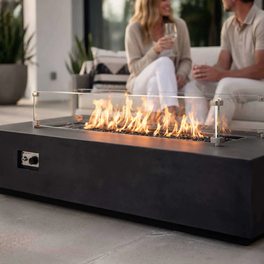 Couple relaxing with drinks behind a lit Flame Havens Refuge Series 68 x 30 x 15-inch rectangular natural gas fire pit in black GFRC concrete. A clear tempered glass wind guard surrounds the flames rising from reflective black fire glass, while the front ignition controls remain visible in the modern patio setting.