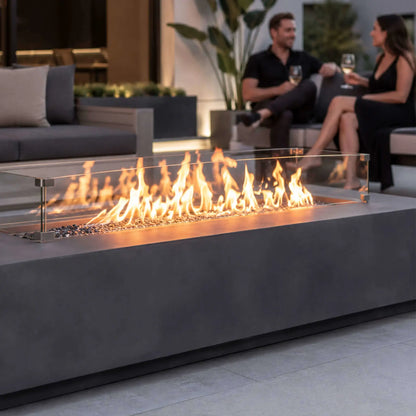 Straight lifestyle view of the Flame Havens Refuge Series 68 x 30 x 15-inch gray GFRC natural gas fire pit with no controls visible. Tall flames burn over tempered black fire glass behind a clear wind guard while a couple enjoys wine on a modern outdoor sofa in the softly lit background.