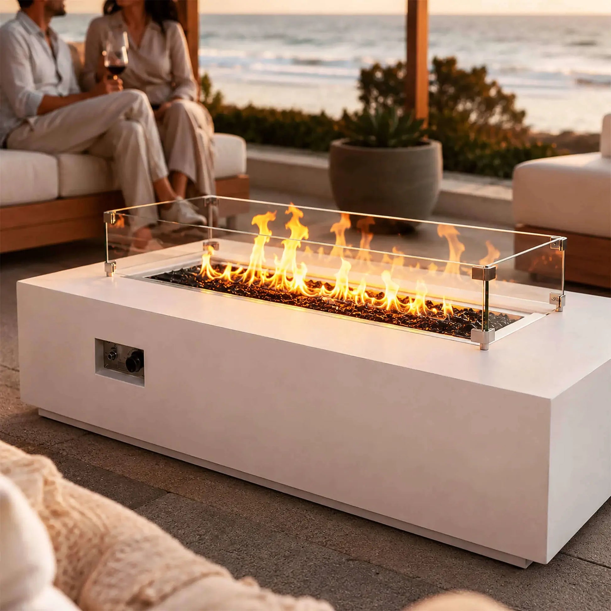 Flame Havens Refuge Series 68 x 30 x 15-inch white GFRC natural gas fire pit glowing on an oceanfront patio at sunset. A clear glass wind guard surrounds the flames and tempered black fire glass, while two adults relax with wine on nearby outdoor seating and the recessed controls remain visible.