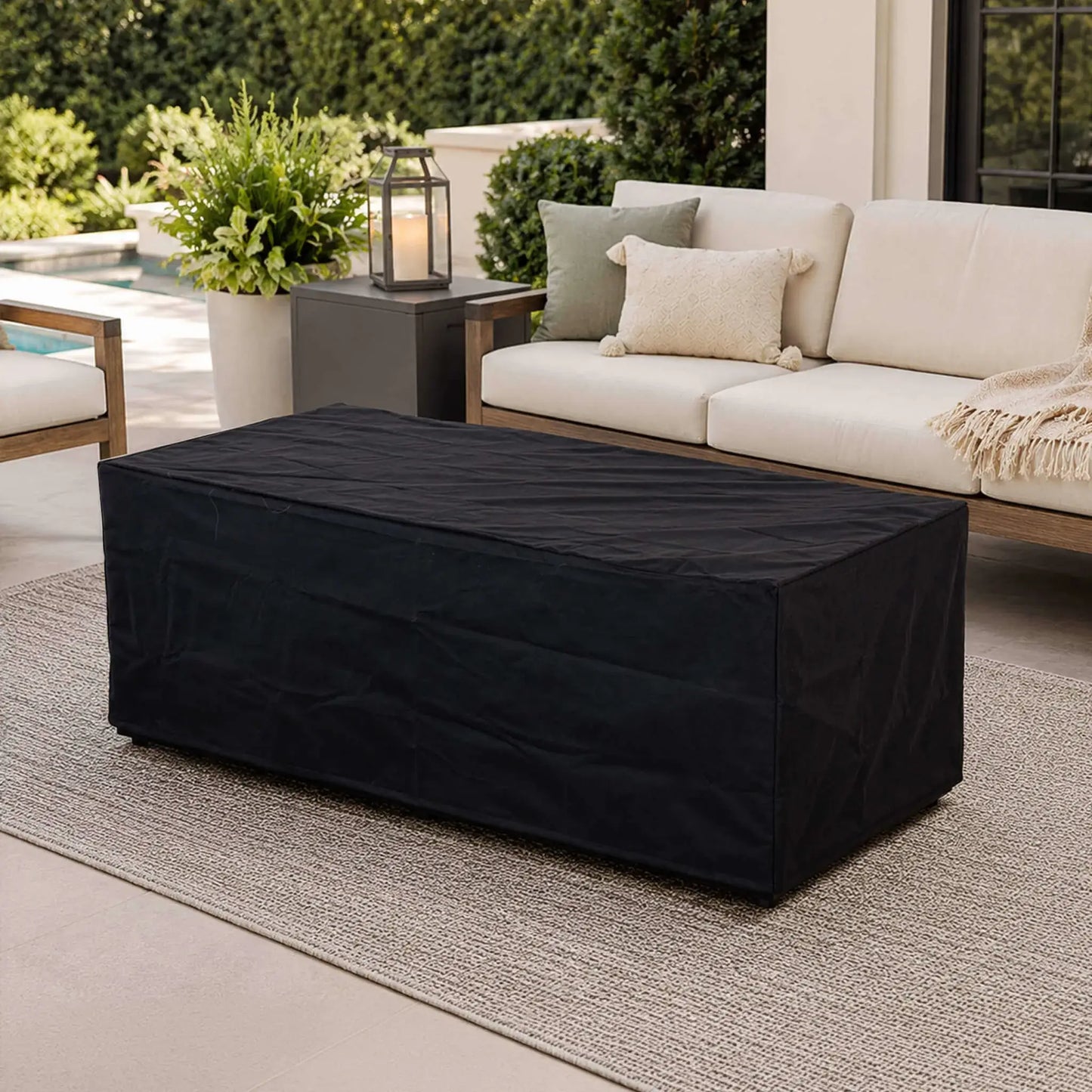 Black canvas fabric cover fitted over the Flame Havens Refuge Series 68 x 30 x 24-inch rectangular GFRC concrete propane fire pit on a refined outdoor patio with cushioned seating, decorative pillows, greenery, a lantern, and a swimming pool in the background.