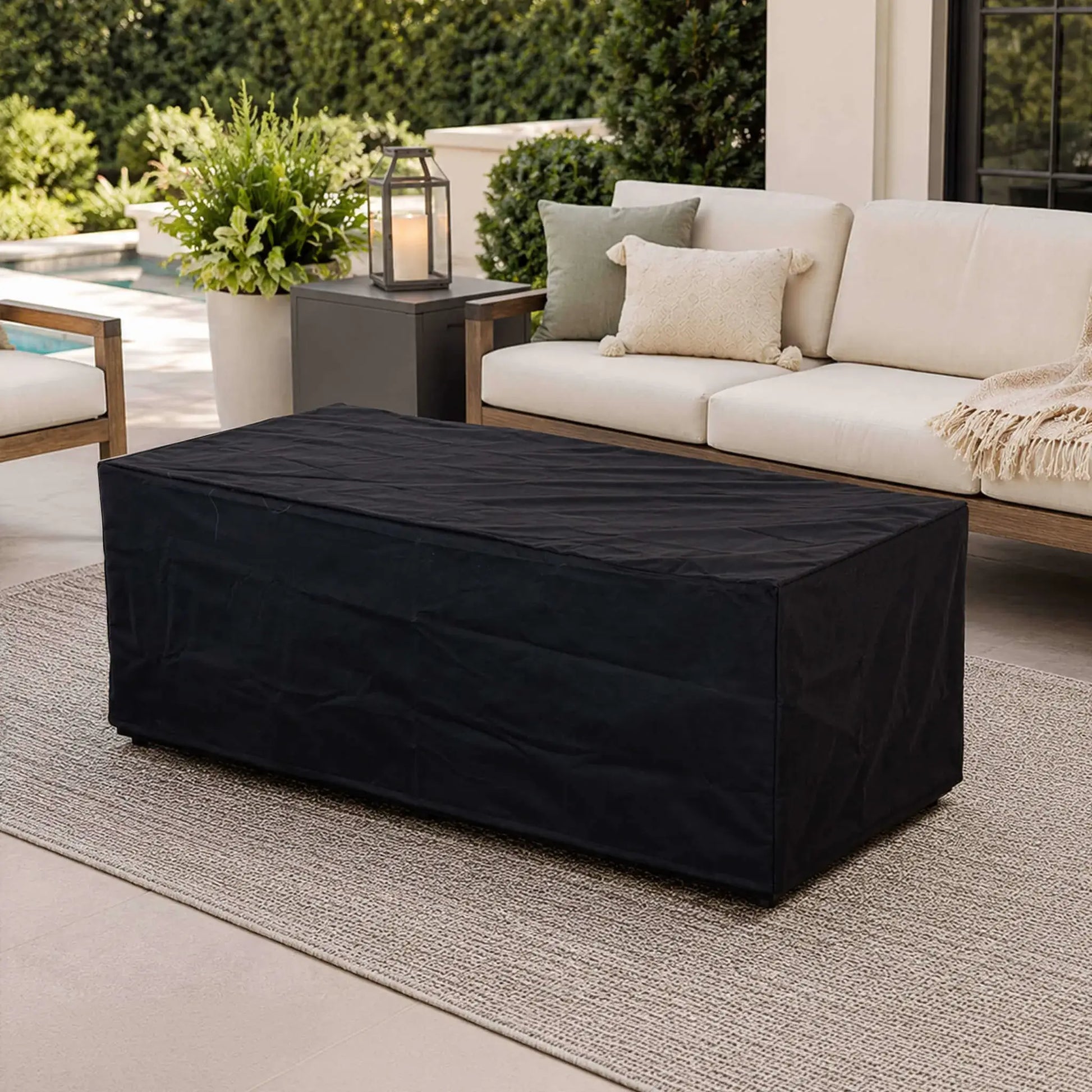 Black canvas fabric cover fitted over the Flame Havens Refuge Series 68 x 30 x 24-inch rectangular GFRC concrete propane fire pit on a refined outdoor patio with cushioned seating, decorative pillows, greenery, a lantern, and a swimming pool in the background.