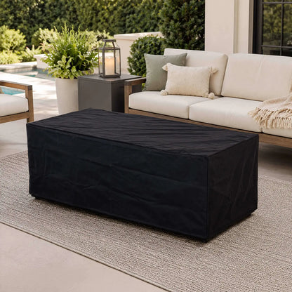 Black canvas fabric cover fitted over the Flame Havens Refuge Series 68 x 30 x 24-inch rectangular GFRC concrete propane fire pit on a refined outdoor patio with cushioned seating, decorative pillows, greenery, a lantern, and a swimming pool in the background.