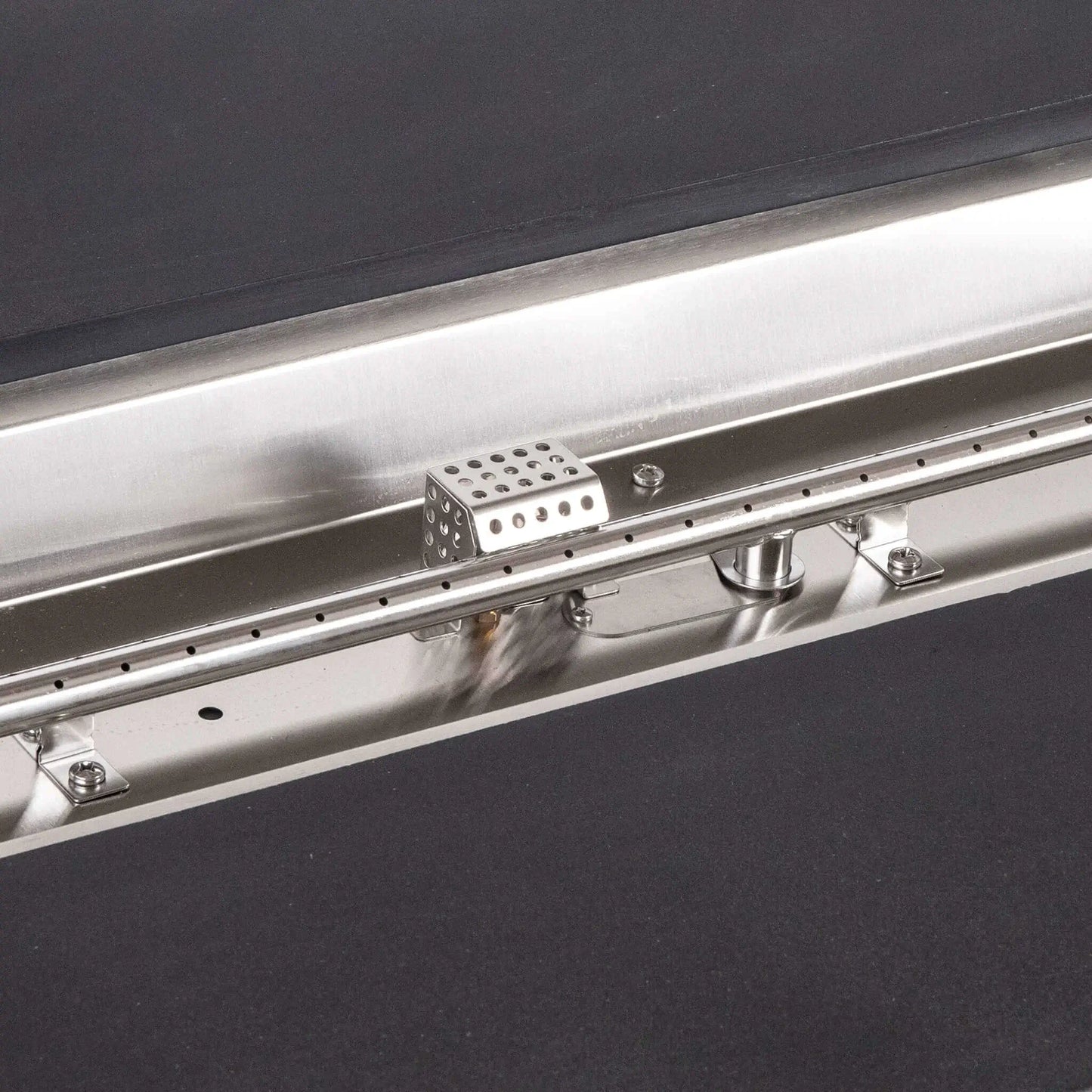 Tight detail of the stainless steel burner and CSA ignition components inside the Flame Havens Refuge Series 68 x 30 x 24-inch black rectangular GFRC concrete propane fire pit, highlighting the linear burner ports, polished pan, hardware, fittings, and perforated ignition cage.