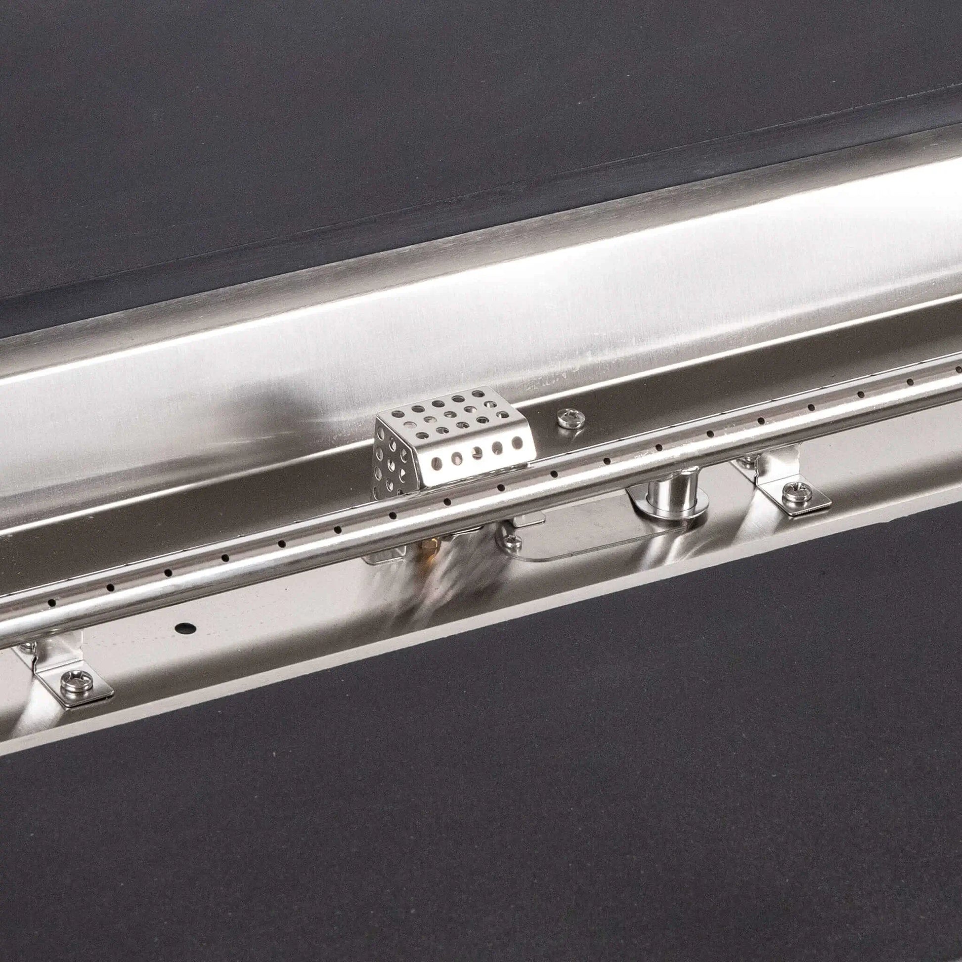 Tight detail of the stainless steel burner and CSA ignition components inside the Flame Havens Refuge Series 68 x 30 x 24-inch black rectangular GFRC concrete propane fire pit, highlighting the linear burner ports, polished pan, hardware, fittings, and perforated ignition cage.