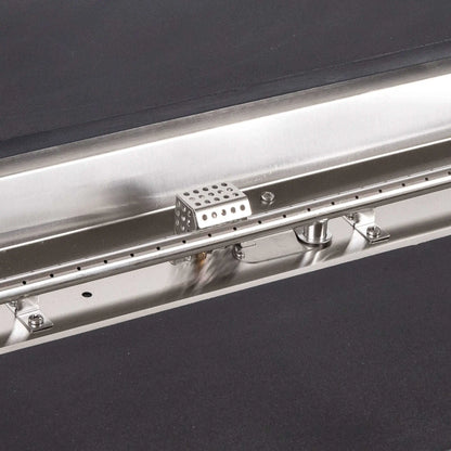 Tight detail of the stainless steel burner and CSA ignition components inside the Flame Havens Refuge Series 68 x 30 x 24-inch black rectangular GFRC concrete propane fire pit, highlighting the linear burner ports, polished pan, hardware, fittings, and perforated ignition cage.
