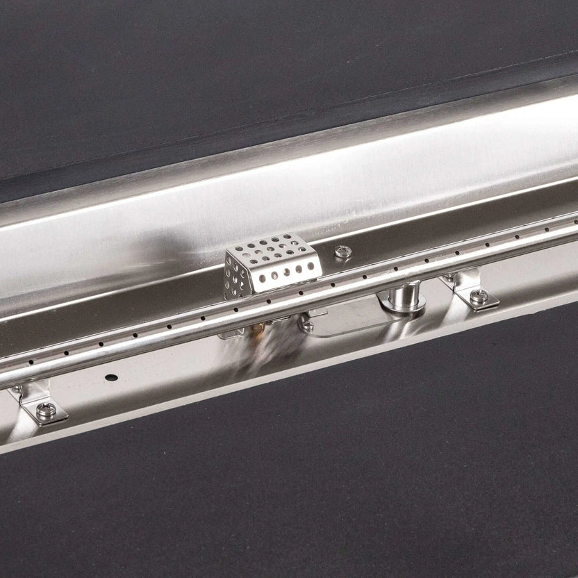 Tight detail of the stainless steel burner and CSA ignition components inside the Flame Havens Refuge Series 68 x 30 x 24-inch black rectangular GFRC concrete propane fire pit, highlighting the linear burner ports, polished pan, hardware, fittings, and perforated ignition cage.