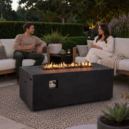 Couple relaxing on outdoor sofas beside a lit Flame Havens Refuge Series 68 x 30 x 24-inch rectangular black GFRC concrete propane fire pit. Flames rise across tempered black fire glass while the front controls and hidden tank drawer blend into the modern patio setting.