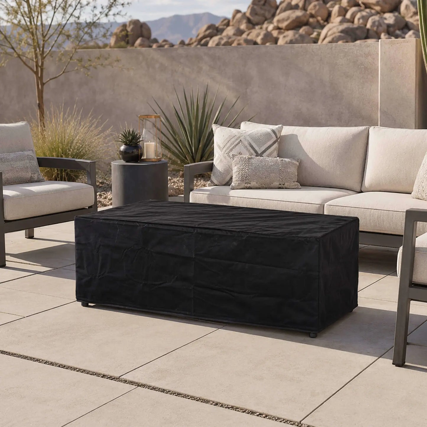 Flame Havens Refuge Series 68 x 30 x 24-inch rectangular gray propane fire pit protected by a fitted black canvas fabric cover on a modern desert patio, surrounded by cushioned outdoor seating, decorative plants, boulders, and mountain views.